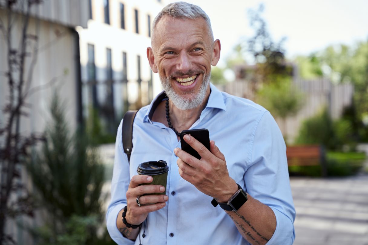 Smiling man walking outside, holding phone and coffee