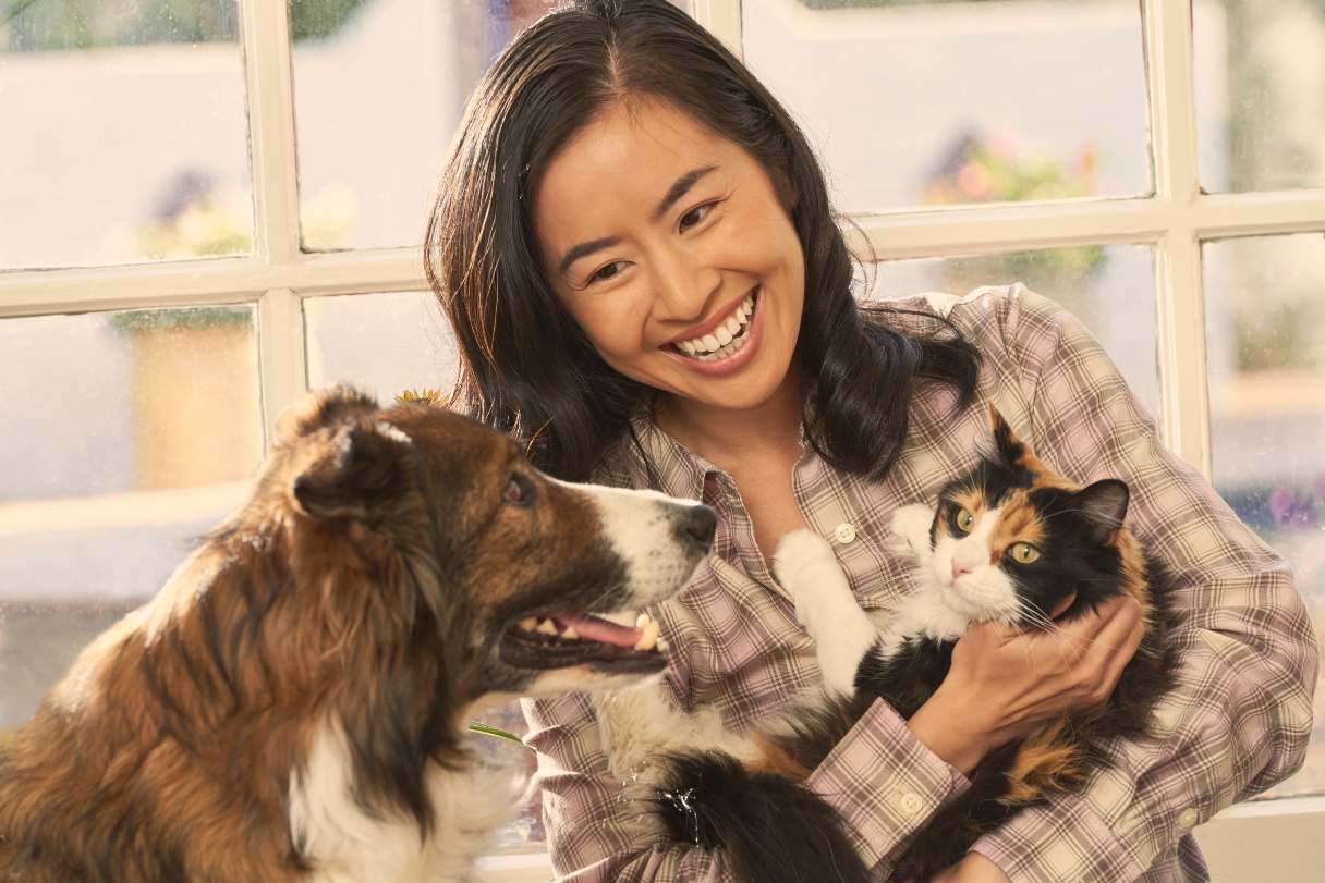 Smiling woman sitting next to a dog, holding a cat