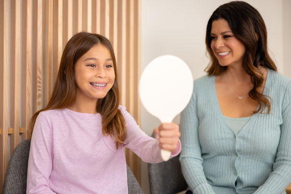 Girl with braces, smiling and looking into hand mirror with woman