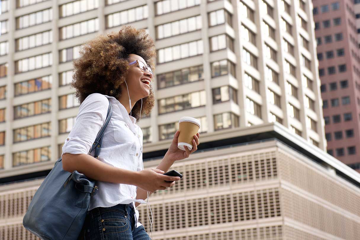 Woman walking next to tall buildings, holding coffee and laughing
